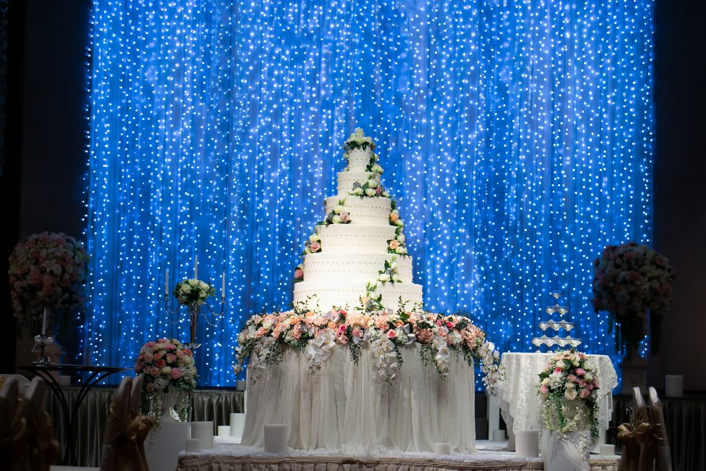 Wedding Cake on the table decorated by beautiful flowers at the stage with beautiful glittering blue light bokeh backdrop in wedding night ceremony. Wedding Ceremony tools and decorations concept.
